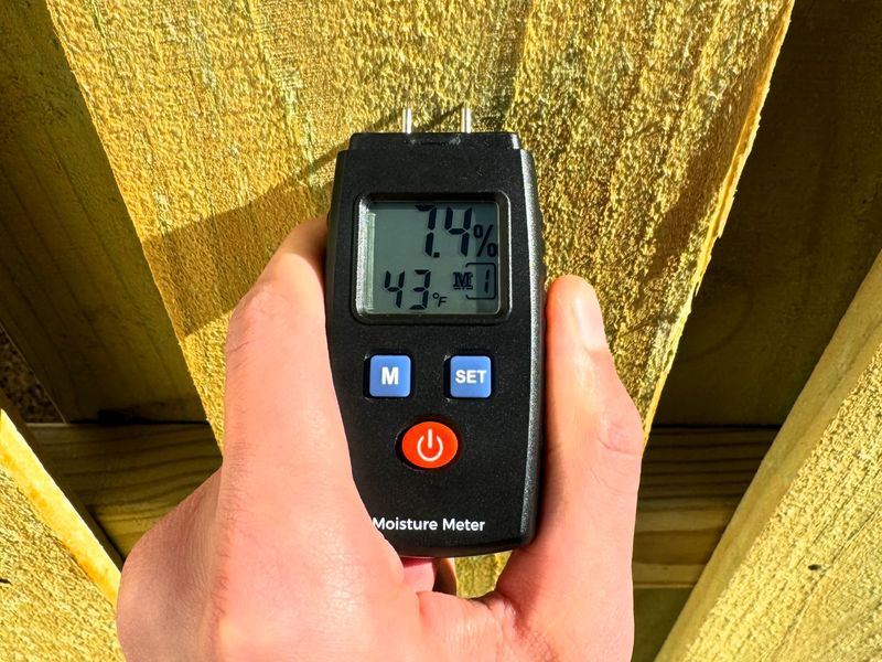 Hand holding a moisture meter on fence boards, reading 7.4 percent