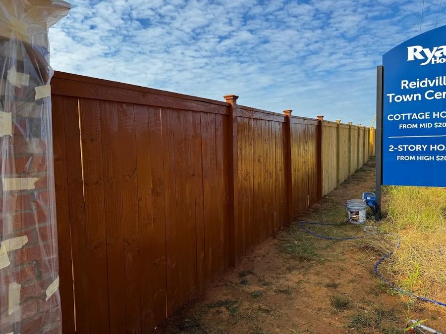 Perimeter privacy fence being stained beside a Ryan Homes sign at Reidville Town Center, stained boards in front and raw boards further down